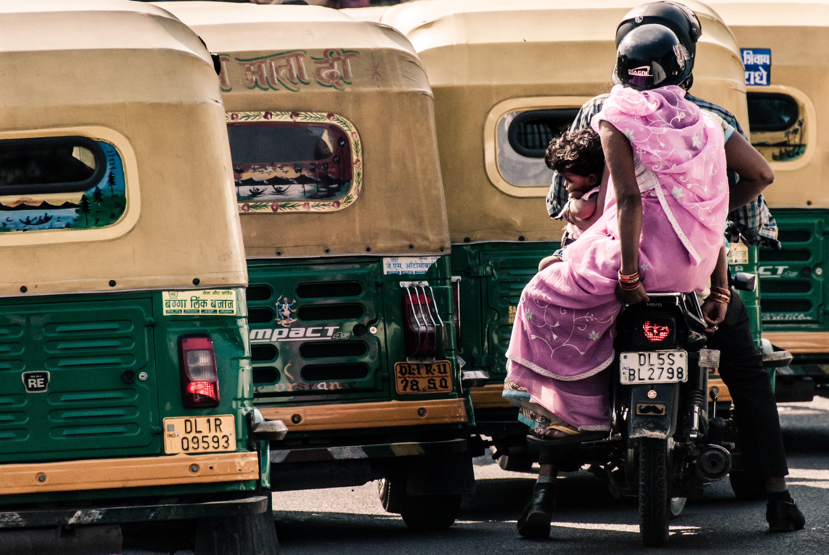 Une passagère en sari rose sur une moto, devant des rickshaws motorisés verts et jaunes