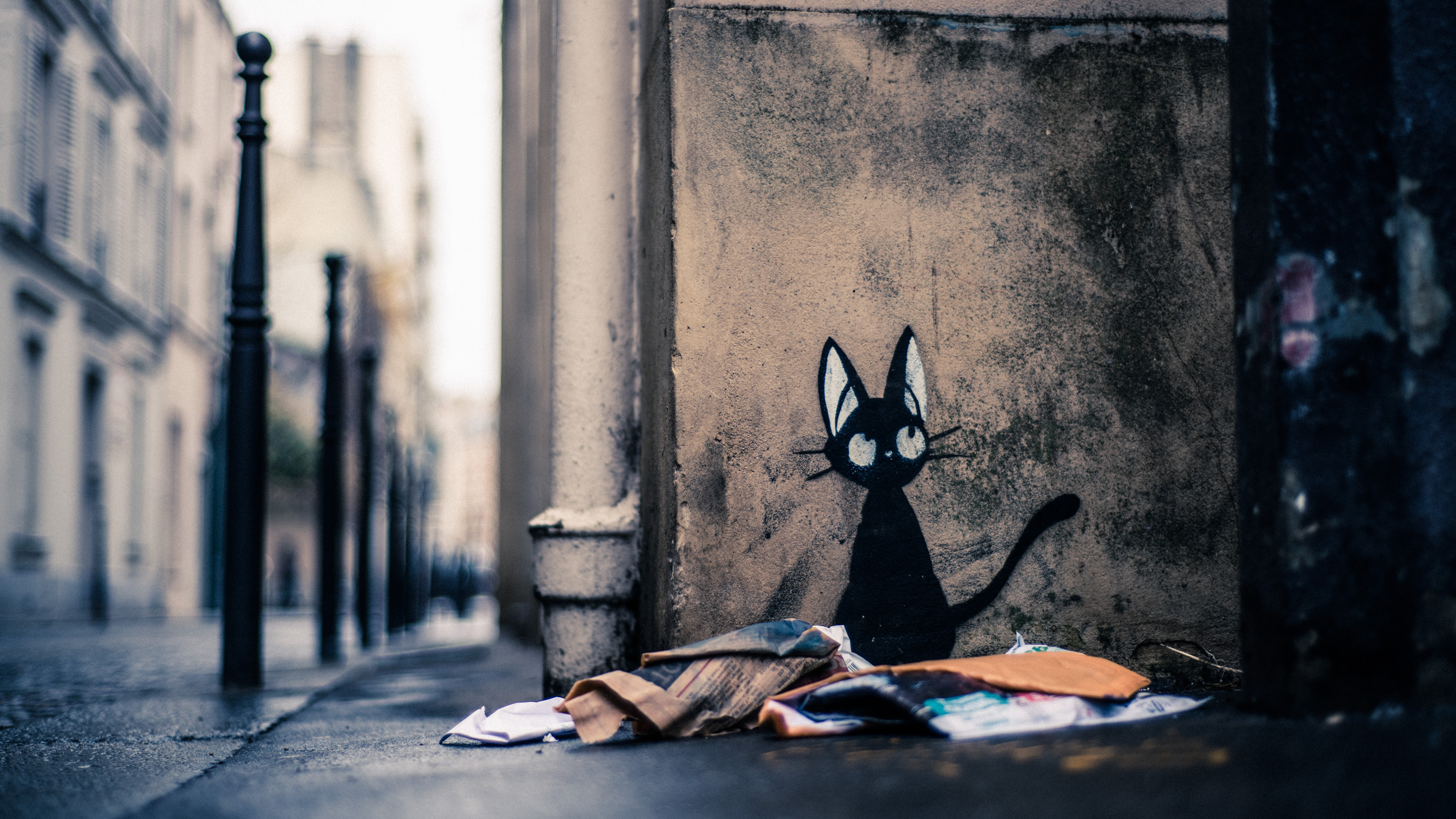 Un chat noir peint sur un mur d'angle, des vêtements en tas au sol, dans une rue étroite